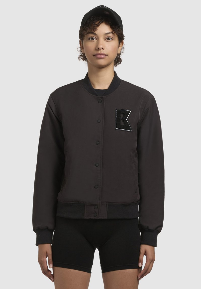 Must jakk musta värvi, ümmarguse kaelusega, nööpidega esiosas ja sooniknæppudega. Vasakul rinnal on tekstuuriga lapiga, millel on 'K'. Minimalistlik disain.