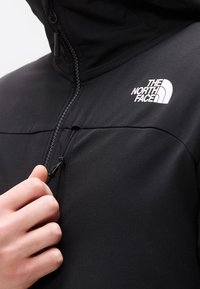 Main d'une personne tirant la fermeture éclair d'une veste noire The North Face avec capuche, montrant le logo sur la partie supérieure gauche de la poitrine.