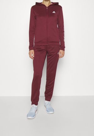 Personne portant un survêtement Adidas bordeaux avec une veste à capuche zippée et un pantalon assorti, accompagné de chaussures de sport bleu clair.