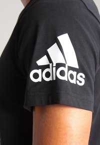 Černé tričko s krátkým rukávem na paži osoby s bílým logem Adidas vytištěným na horním rukávu.