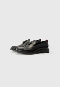 Black leather shoes featuring brogue detailing, a rounded toe, and a small heel. Accents include a decorative strap with a buckle.