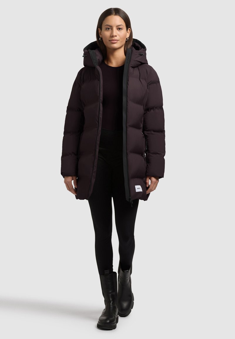 Chaqueta de plumas negra con capucha, que presenta una cremallera frontal completa, bolsillos laterales y un parche con logo blanco en la parte inferior derecha. Textura suave.