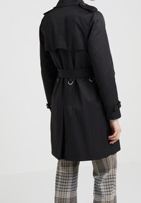 Czarny trench coat z paskiem w talii, podwójnymi guzikami oraz zapinanymi na guziki naramiennikami. Posiada rozcięcie z tyłu i teksturowany materiał.