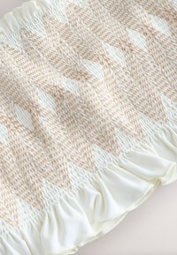 Balts audums ar krēmkrāsas saspesto izšuvumu, kas veido ģeometriskus rakstus un savāktu volānu malu.