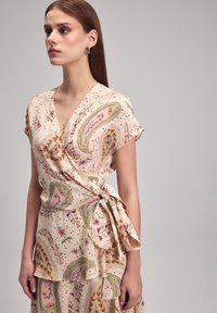 Wickelbluse in hellem Creme mit Paisley- und Blumenmustern in Grün, Pink und Rot. Kurzärmelig, mit gebundenem Taillengürtel, aus weichem Stoff, mit geschichtetem Design.
