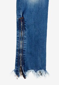 Blå denimjeans med fransade kanter och exponerad dragkedjedetalj längs sidan. Har kontrasterande sömmar och en slim fit-design.