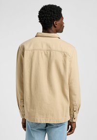 Chaqueta de algodón beige con cuello puntiagudo, puños abotonados y una yugo trasero con un pequeño logotipo. La textura parece suave y duradera.