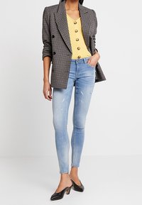 Rutig dubbelknäppt kavaj, gul skjorta, slitna ljusblå skinny jeans och svarta klackskor med spetsiga tår. Enkla färger och texturer.