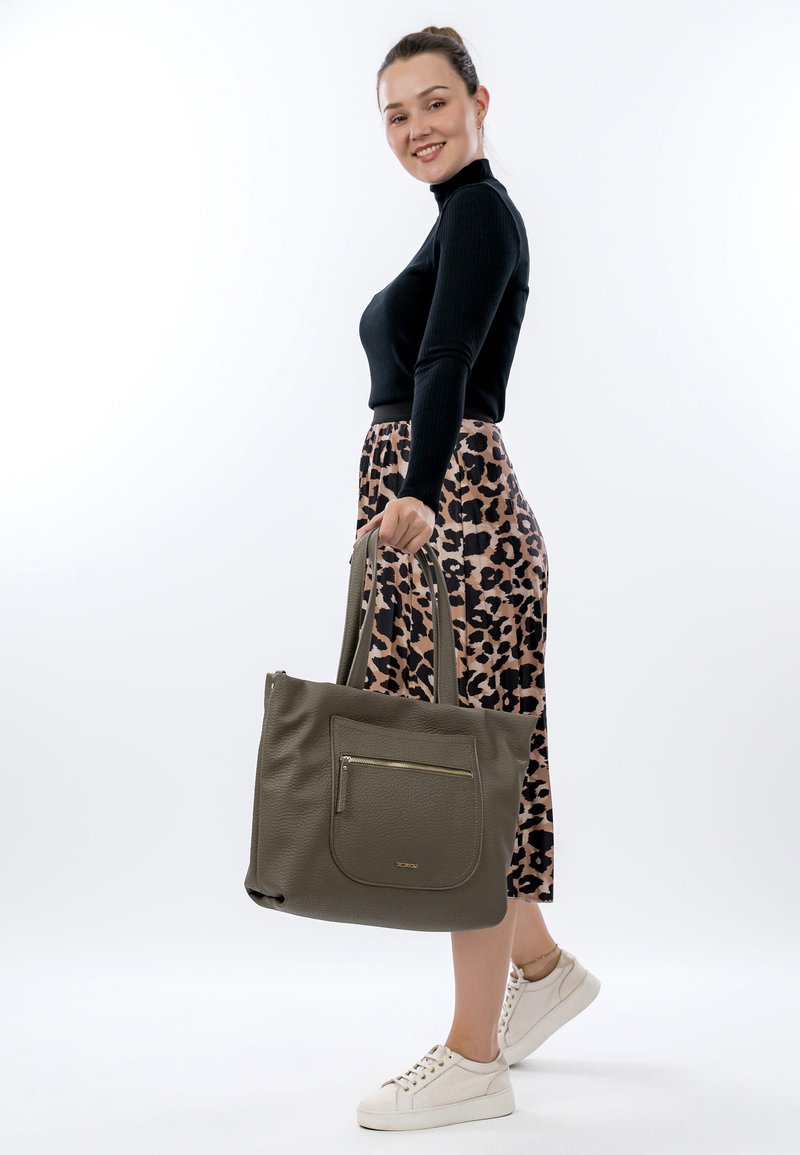 Olive green tote bag with textured surface, large front pocket, and gold zipper accents. Worn with a black top and leopard print skirt.