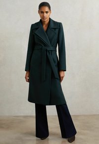 Naine seisab, seljas tume roheline vööga mantel, mustade laiade pükste ja tumedate kingadega, lihtsa heleda tausta ees.