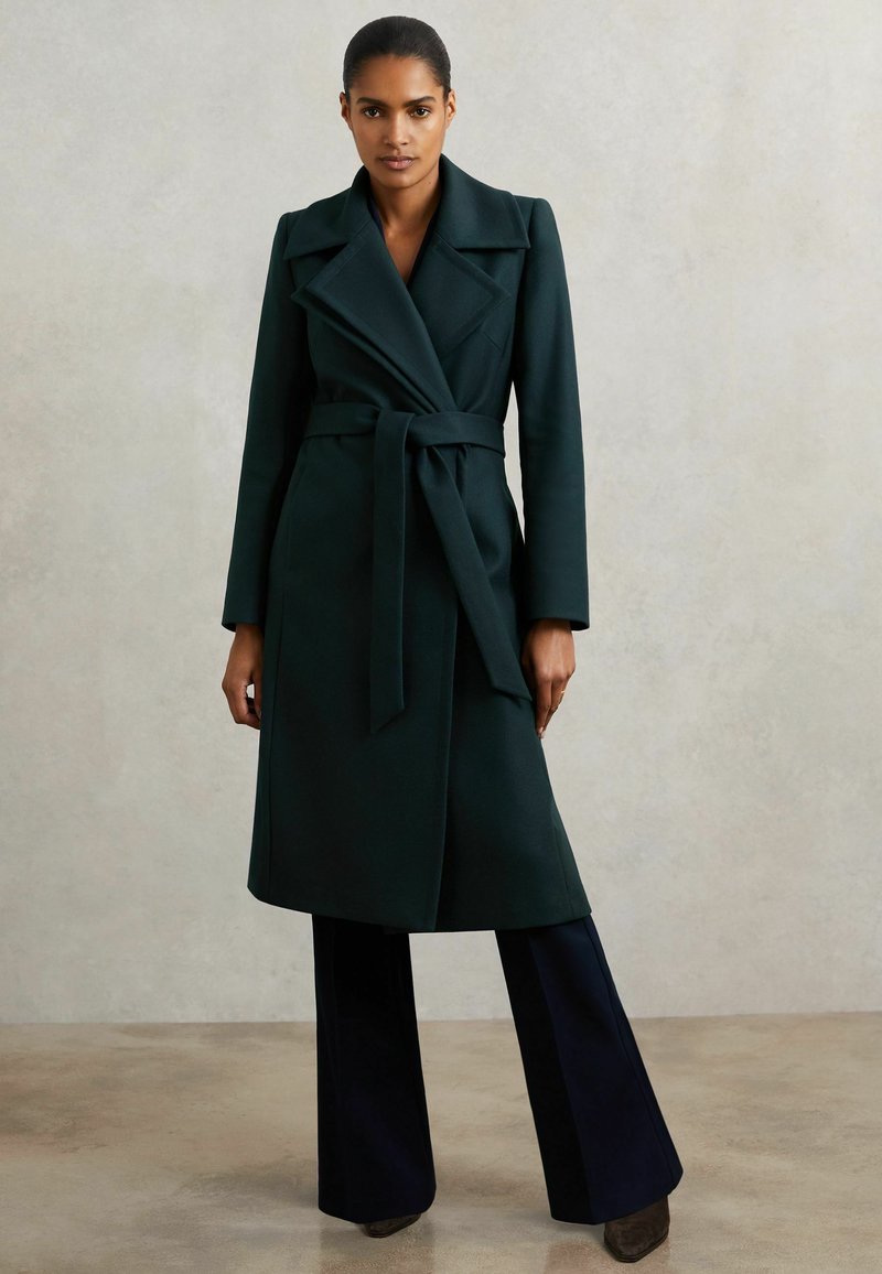 Naine seisab, seljas tume roheline vööga mantel, mustade laiade pükste ja tumedate kingadega, lihtsa heleda tausta ees.