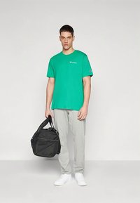 Bărbat îmbrăcat cu un tricou verde Champion, pantaloni de trening gri deschis, adidași albi, ținând o geantă de sală neagră pe un fundal simplu.