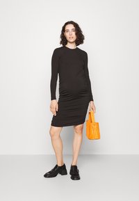 Robe de maternité noire à manches longues, en maille côtelée, avec des côtés froncés, longueur genoux. Associée à un sac à main en suède orange et des mocassins noirs.