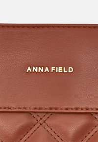 Portefeuille en cuir marron texturé avec un motif en losanges, présentant un logo doré "ANNA FIELD". Détail de couture le long des bords.