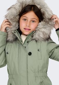 Roheline pehme talvejope, millel on eemaldatav hall karvane kapuuts, lukuga ja nööbiga sulgemine ning elastne vöökoht, et paremini sobida.