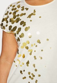 Vit kortärmad skjorta med spridda metalliska guldleopardfläckar på framsidan och axeln, bärs av en person.