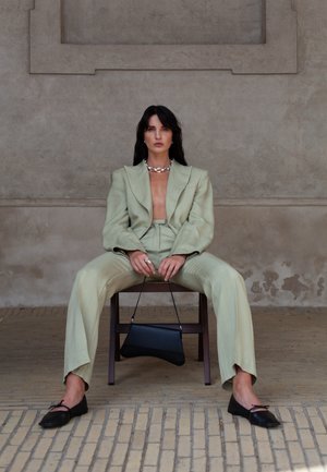 Femme aux longs cheveux foncés assise sur une chaise en bois contre un mur texturé, portant un costume vert clair, des ballerines noires, un collier en argent, tenant un sac à main noir.