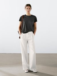 Sort kortærmet t-shirt parret med brede bukser med hvide pinstriber. Modellen står med hænderne i lommerne og holder en brun flettet taske.
