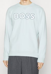 Ljusblå sweatshirt i bomullsblandning, med en stor "BOSS"-logotyp i mörkblått över bröstet, samt ribbade mudd och nederkant.