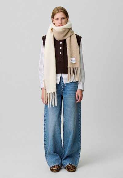 Gilet en tricot marron avec écharpe beige et crème ; jean denim bleu ample ; chaussures à imprimé animal ; rayures verticales sur la chemise en dessous.