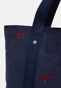 Sac fourre-tout en velours côtelé marine avec logo rouge brodé ; dispose d'une épaisse sangle et d'un détail de bouton-pression. Surface texturée avec des rayures verticales.