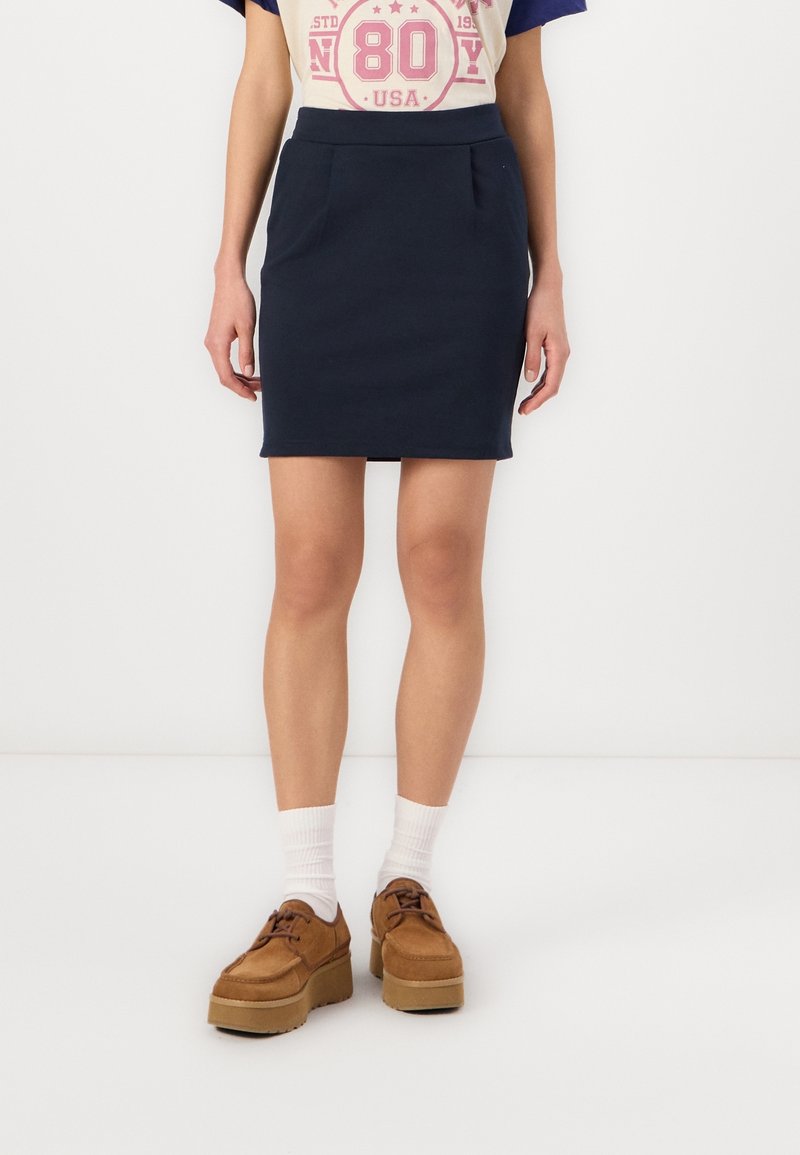 Mörkblå figursydd kjol med sidofickor, tillsammans med en gräddfärgad och marinblå grafisk t-shirt. Bruna platåskor och vita strumpor fulländar outfiten.