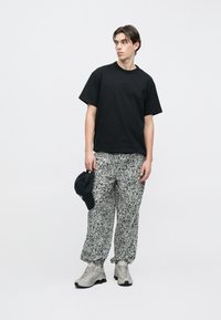 Młody mężczyzna ubrany w czarną koszulkę Nike, wzorzyste luźne spodnie, szare buty sportowe Nike, trzymający czarną czapkę, stojący na białym tle.