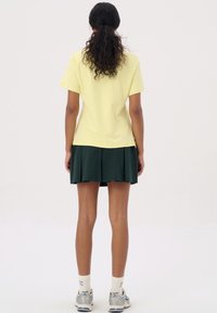 Geel kort shirt met korte mouwen, gecombineerd met een donkergroene geplooide rok. Model heeft donker, krullend haar dat naar achteren is vastgebonden. Sneakers en witte sokken maken de outfit compleet.