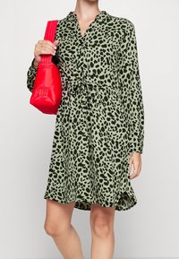 Grøn langærmet kjole med sorte leopardpletter, smalt indsnøret i taljen, sat sammen med en rød håndtaske med en struktureret rem.