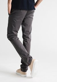 Mężczyzna stoi z nogami skrzyżowanymi, ubrany w ciemnoszare spodnie, beżowe trampki i czarną koszulę, trzymając ręce w kieszeniach.