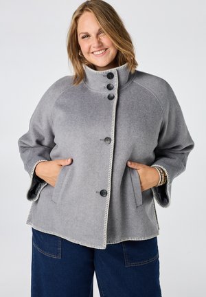 Femme souriante portant un manteau en laine gris boutonné avec des poches et un pantalon bleu foncé, debout devant un fond clair uni.