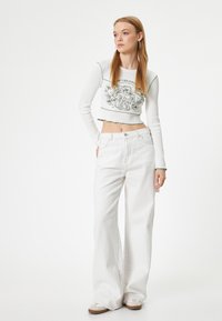 Witte cropped longsleeve top met groene accentstiksel en grafische print, gecombineerd met hoge taille, wijdvallende witte jeans en lichte schoenen.