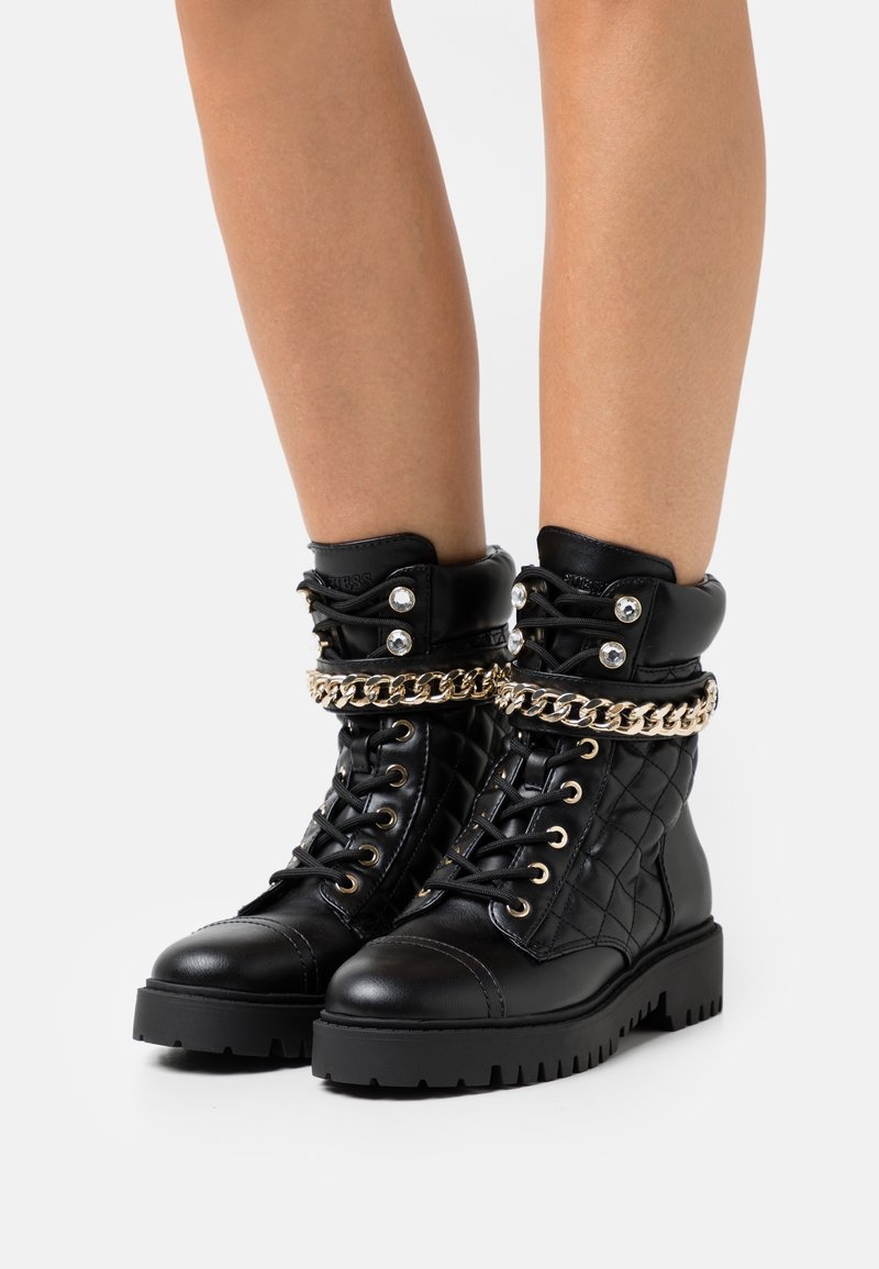 Botas de combate de cuero negro con textura acolchada, diseño de cordones y un acento de cadena dorada alrededor del tobillo. Suela gruesa para mayor soporte.