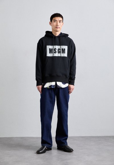 Schwarzer Kapuzenpullover mit breitem weißem Text "MSGM", Fronttasche, getragen über einem hellen Hemd, kombiniert mit weiten dunkelblauen Jeans und schwarzen Schuhen.