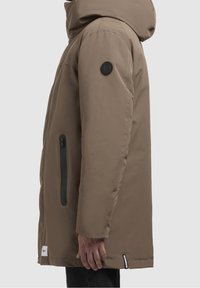 Parka impermeable marrón con capucha, con un parche de logo negro en la manga y un bolsillo lateral con cremallera. Textura suave y lisa.