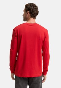 Mann mit kurzen braunen Haaren, der sich abwendet, trägt ein schlichtes rotes Langarmshirt und dunkelgraue Jeans vor einem weißen Hintergrund.