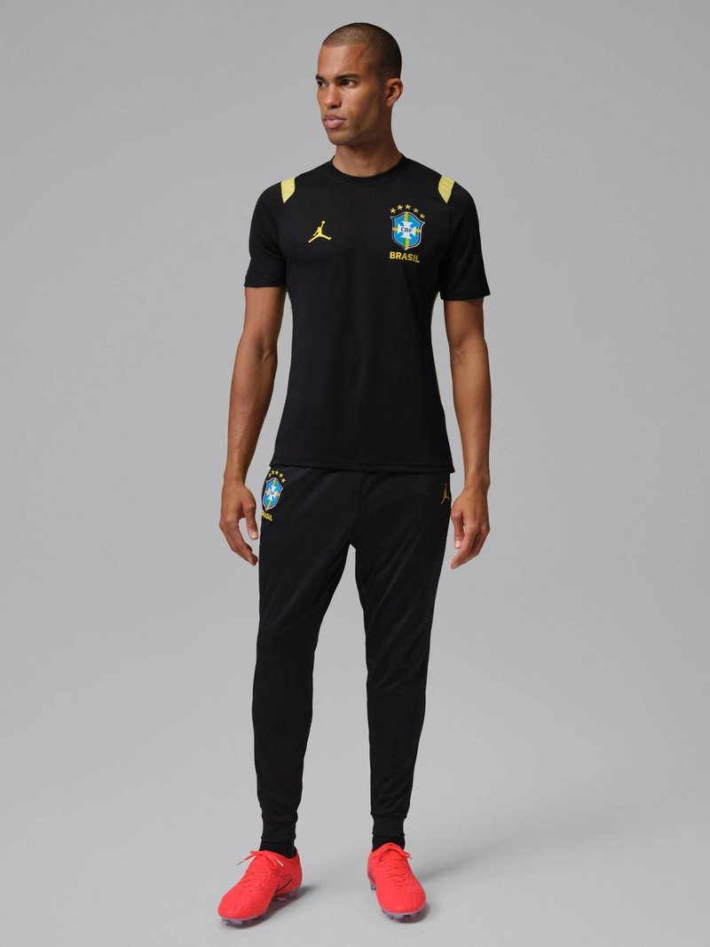 Man staat in een zwart Braziliaans voetbaltrainingspak met gele logo's en felrode voetbalschoenen tegen een grijze achtergrond.