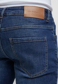 Lähedane plaan inimesest, kes kannab tumehallidest denimpükse, millel on oranž õmblusja hall nahast silt "ZV PREMIUM" vööl.