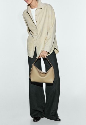 Torebka ze skóry koniakowej o zakrzywionym kształcie i krótkim uchwycie, trzymana przez osobę w kremowej, oversize'owej koszuli i ciemnych spodniach o szerokich nogawkach.