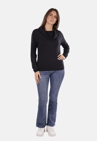 Maglione nero con colletto a cowl e maniche lunghe, indossato sopra una maglietta nera, abbinato a jeans svasati blu e sneakers bianche. Tessuto morbido, vestibilità casual.