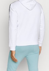 Sweat à capuche blanc avec une poche kangourou, doté de bandes latérales noires. Porté avec un pantalon de jogging bleu sarcelle clair, affichant la vue de dos et les détails de la capuche.