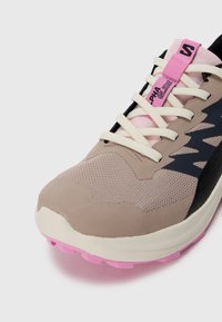 Scarpa da ginnastica beige e nera con una tomaia in rete e materiale sintetico, lacci rosa e una suola rosa texturizzata. Presenta un logo prominente e accenti di design.
