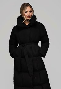 Žena s blond vlasy, oblečená ve velkých zlatých náušnicích a černém péřovém kabátu s vysokým límcem, má ruce v kapsách, na šedém pozadí.