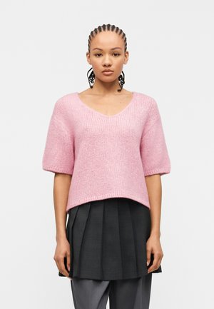 Fonott hajú nő rózsaszín, bolyhos, rövid ujjú pulóvert, fekete pliszírozott szoknyát és szürke nadrágot visel, előre nézve, fehér háttér előtt.