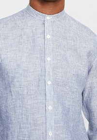 Camicia a righe blu chiaro con colletto alla cinese, realizzata in un tessuto traspirante. Presenta piccoli bottoni bianchi lungo la parte anteriore.