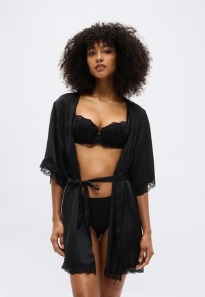 Γυναίκα με σγουρά μαλλιά που φοράει μαύρο δαντελωτό σουτιέν, ταιριαστό εσώρουχο και μαύρη σατέν ρόμπα με δαντελένιο τελείωμα δεμένη στη μέση, όρθια μπροστά σε λευκό φόντο.