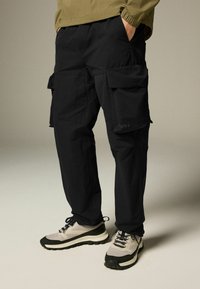 Pantaloni cargo negri cu multiple buzunare, talie elastică și croială lejeră, asortați cu adidași gri și negri cu tălpi texturate.