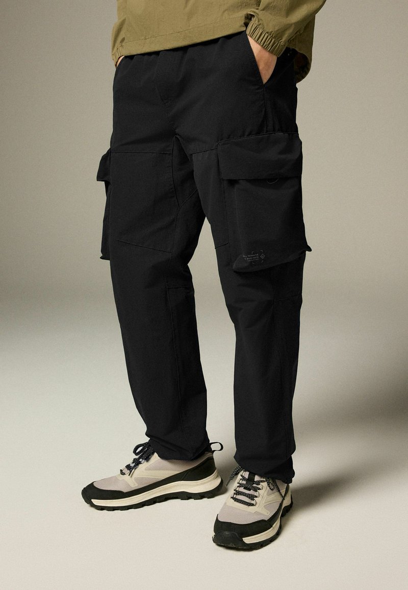 Pantaloni cargo negri cu multiple buzunare, talie elastică și croială lejeră, asortați cu adidași gri și negri cu tălpi texturate.