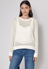 Lekki sweter z kremowej wełny o fakturowanym, ażurowym wzorze. Długie rękawy, okrągły dekolt i ściągacze przy mankietach. W połączeniu z niebieskimi dżinsami.