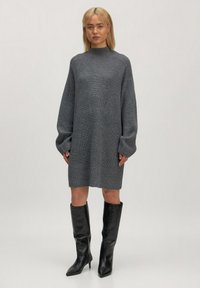Graues Strickkleid mit langen Ärmeln, hohem Kragen und gerippter Textur, kombiniert mit knielangen schwarzen Stiefeln mit Absatz.